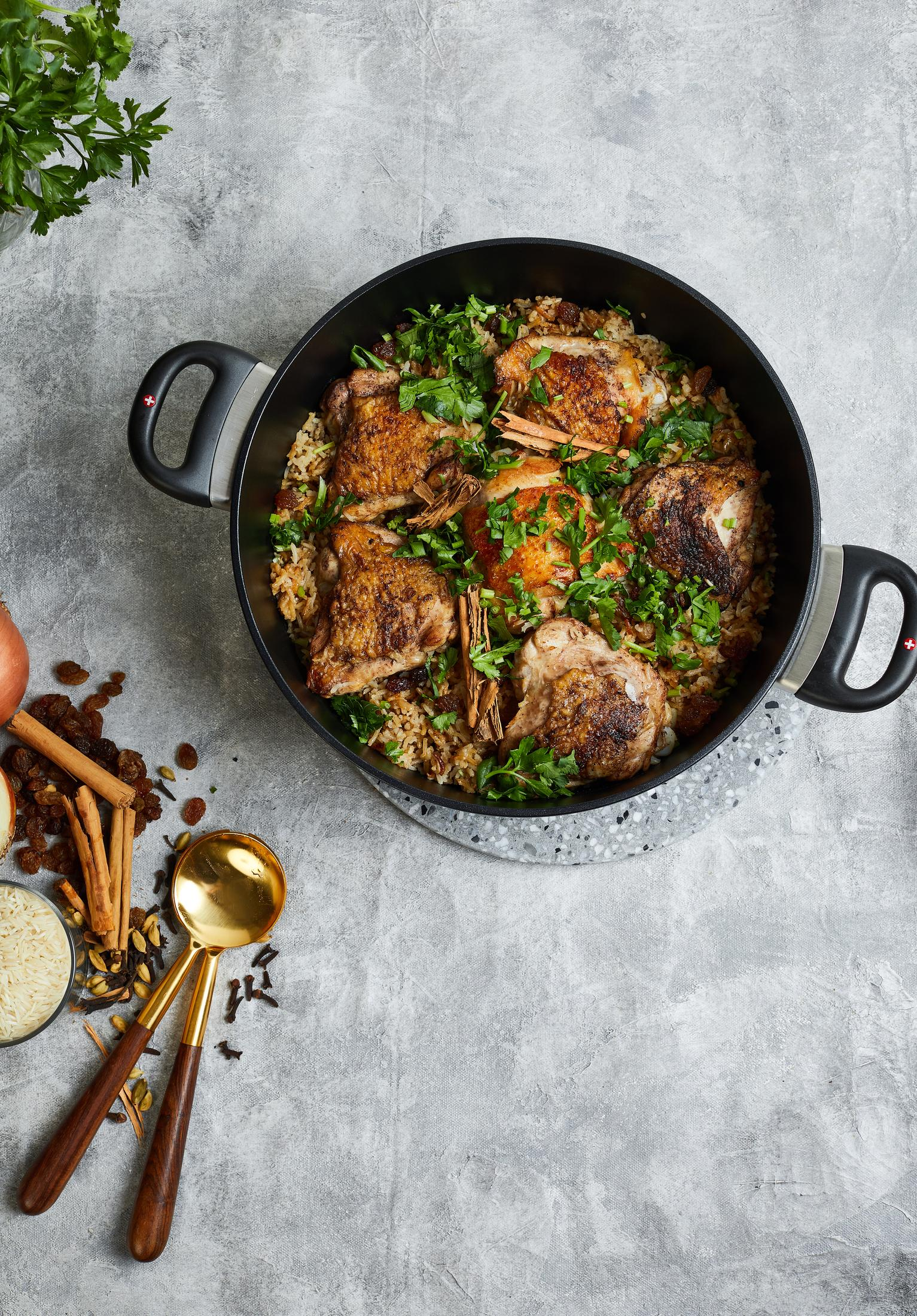 Ein Topf mit Hühnchen, Reis und Gewürzen, daneben Rosinen, Zimt und Löffel.