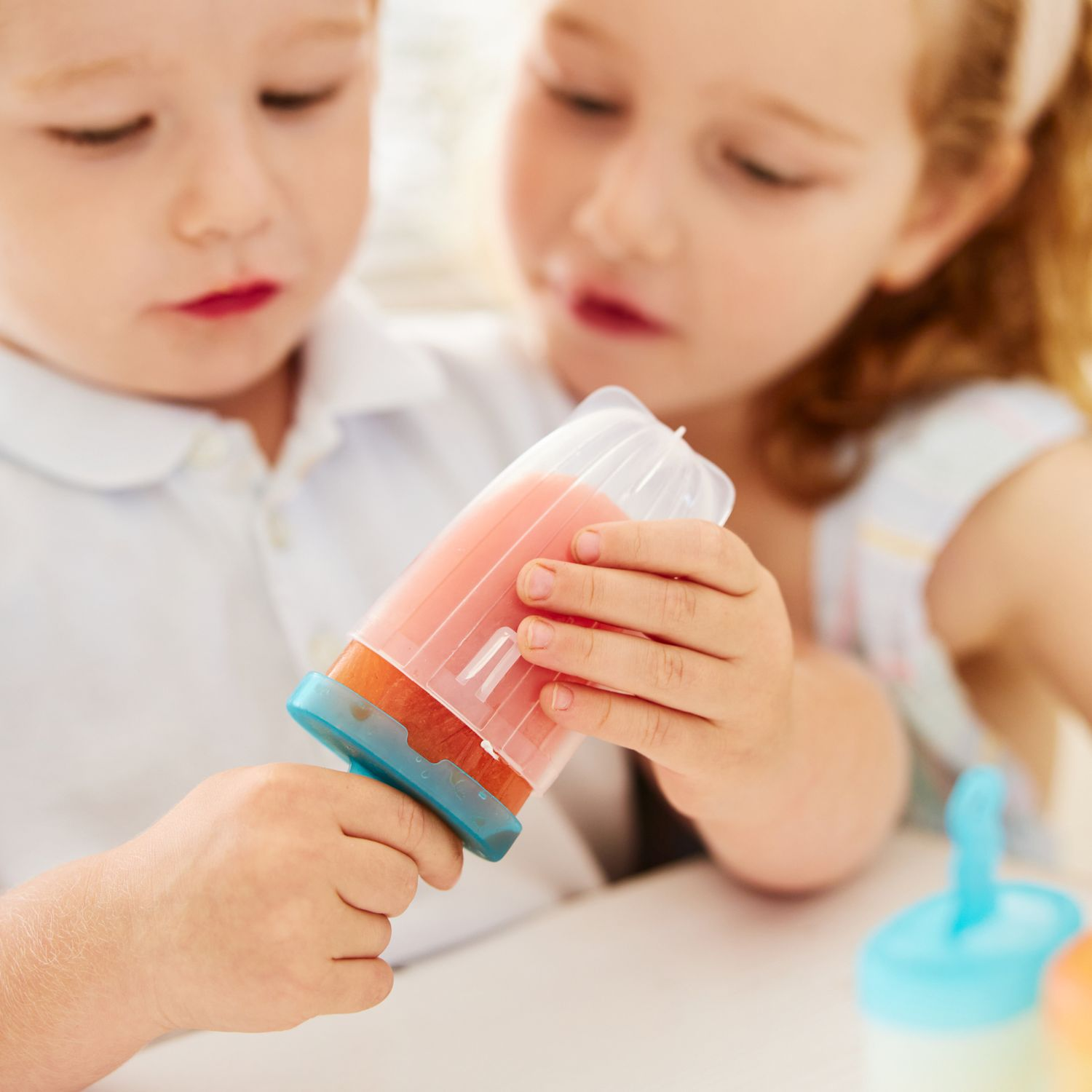 Due bambini tengono un ghiacciolo rosa con manico blu. Si vede un altro ghiacciolo.