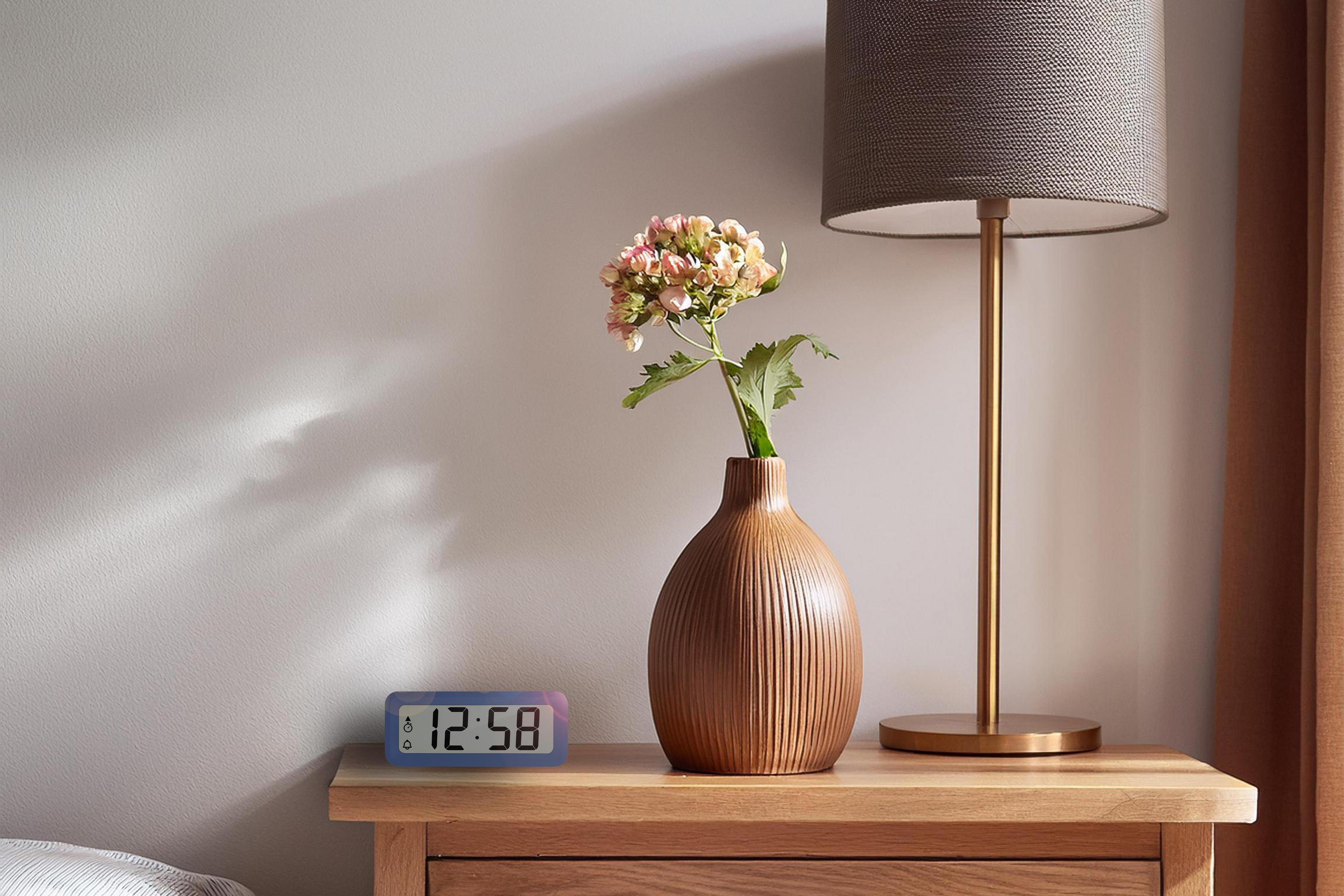Ein digitaler Wecker zeigt 12:58 Uhr. Daneben eine Vase mit Blumen und eine Lampe.