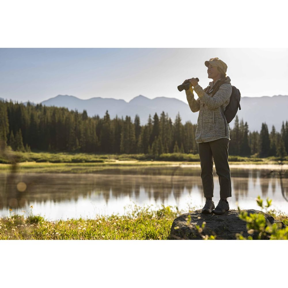 Kaya üzerinde dürbünlü kadın, göl, orman ve dağlar arka planda.