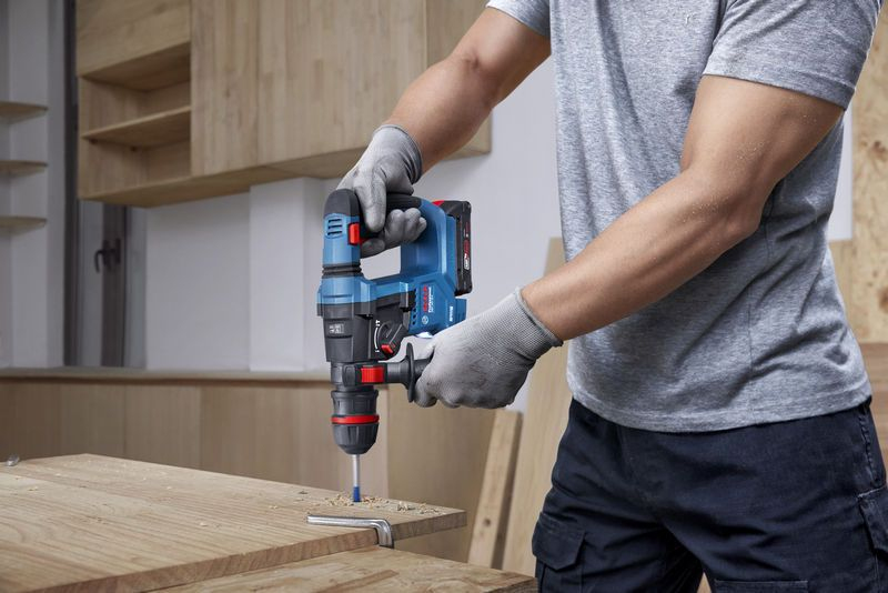 Person in grauem Hemd und Handschuhen benutzt blauen Bohrhammer an Holz.