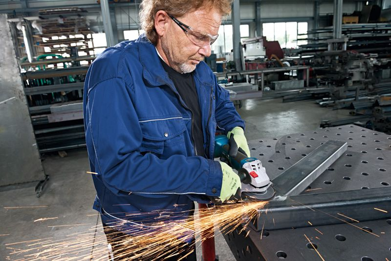 Mann in blauer Jacke und Schutzbrille schleift Metall, Funken fliegen in Werkstatt.