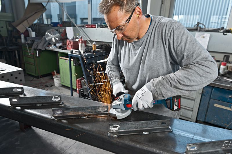 Mann schleift Metall mit Winkelschleifer; Funken sprühen. Werkstatt im Hintergrund.