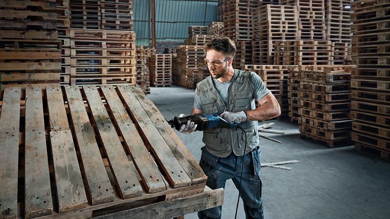 Un uomo con occhiali protettivi e guanti lavora su un pallet di legno con un attrezzo in un magazzino pieno di pallet.