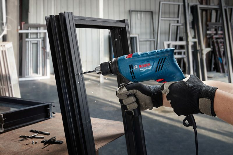 Mavi bir Bosch matkap, yakındaki aletlerle siyah metal profile delik açıyor.