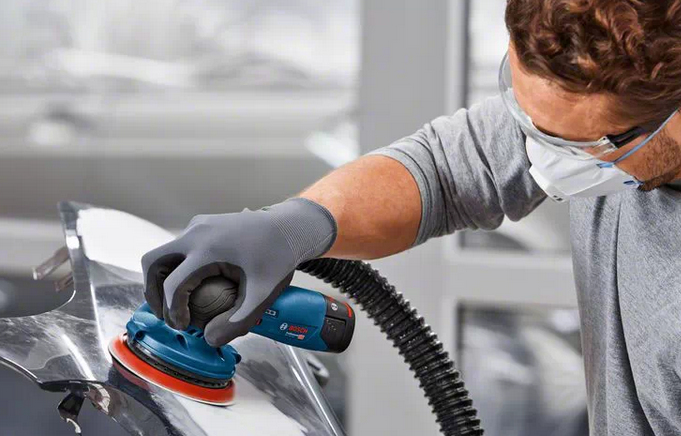 Eine Person in Schutzkleidung schleift eine Autokarosserie mit einem blauen Exzenterschleifer.