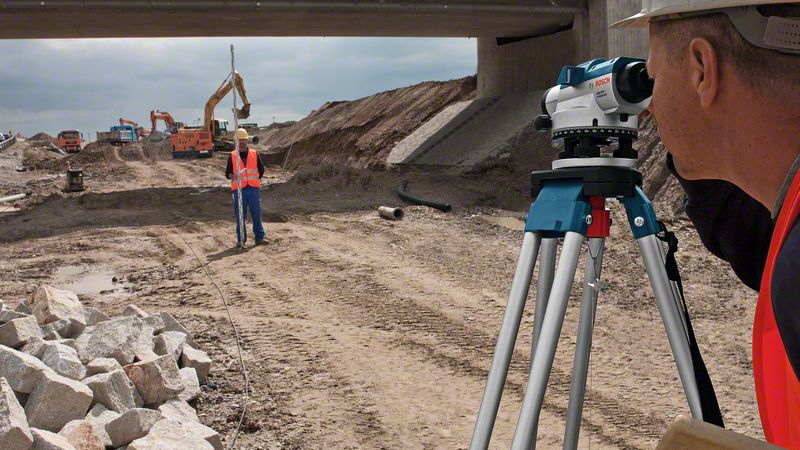 Ein Vermesser blickt durch ein Nivelliergerät auf einer Baustelle mit Baggern und Lastwagen im Hintergrund.