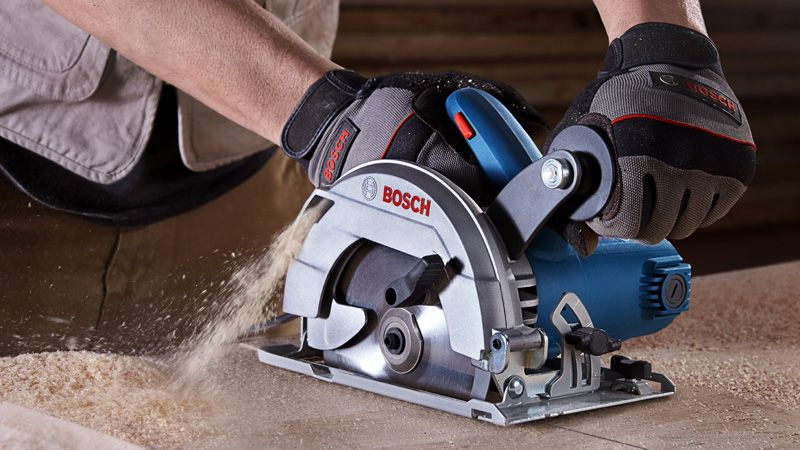 Bosch dairesel testere ahşap kesiyor, talaş üretiyor. Ellerde eldiven var.