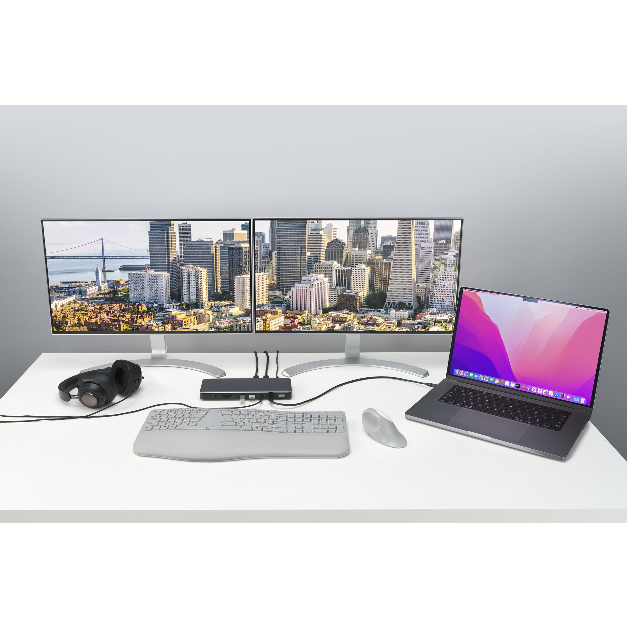 A desk with dual monitors displaying a cityscape, laptop, keyboard, mouse, and headphones.