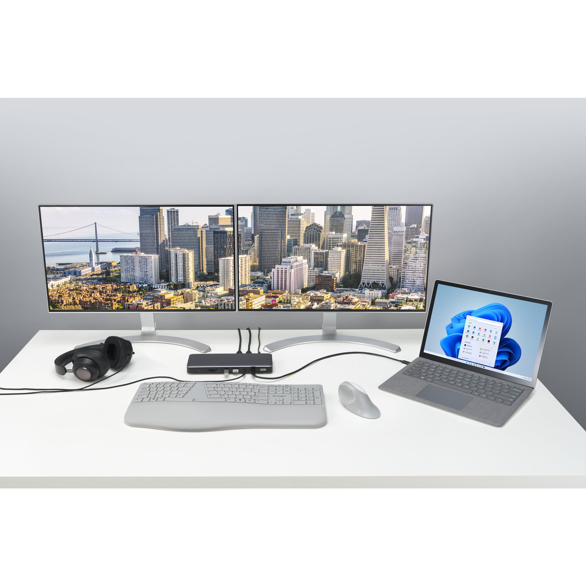 Two monitors display a cityscape. A laptop, keyboard, mouse, and headphones rest on a desk.