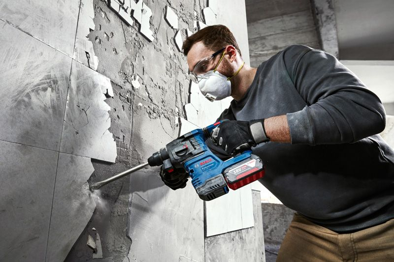 Mann mit Schutzbrille und Maske bricht Fliesen mit Bohrhammer ab.