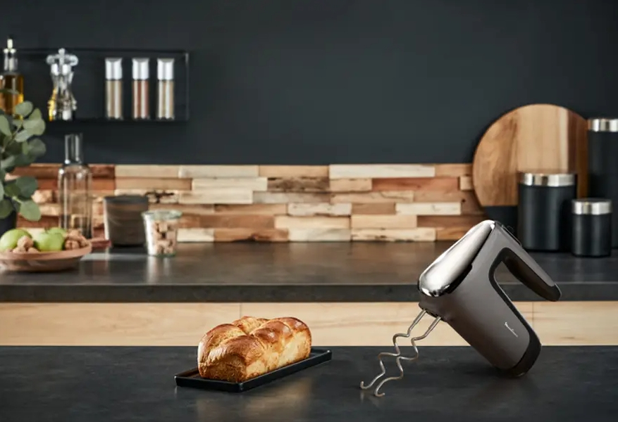 Un batidor de mano junto a un pan sobre una encimera de cocina oscura.