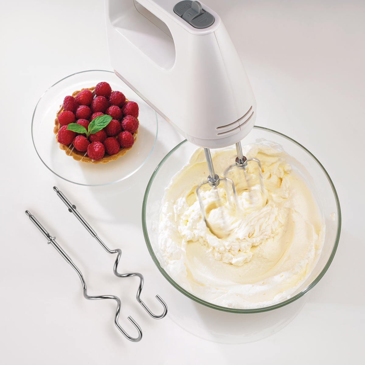 Ein weißer Handmixer schlägt Sahne in einer Schüssel neben einer Himbeertarte.