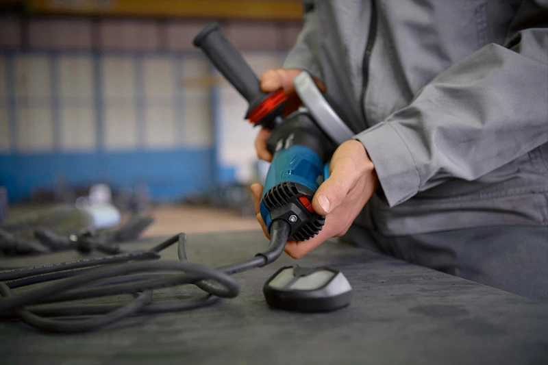 Person in grauer Arbeitskleidung hält blaues Elektrowerkzeug mit Kabel.