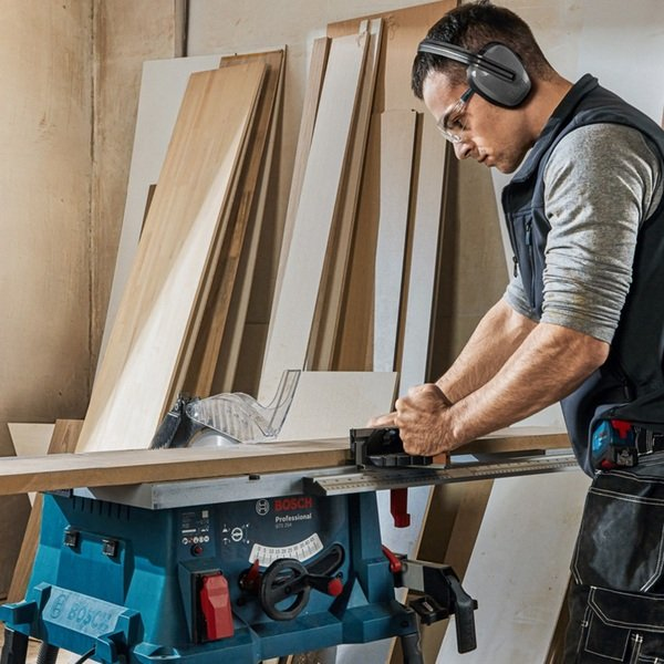 Mann schneidet Holz mit Tischkreissäge, trägt Gehörschutz und Schutzbrille.