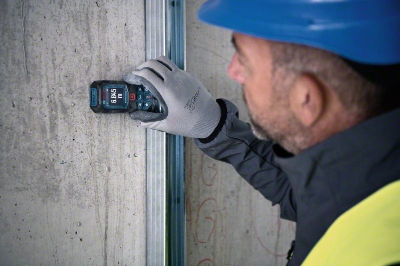 Mann mit blauer Kappe benutzt digitales Messgerät an Betonwand neben Metallbolzen.