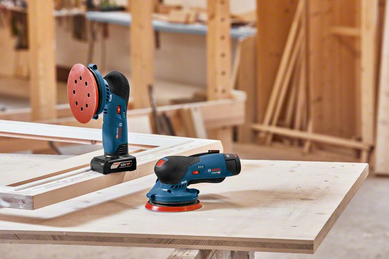 Dos lijadoras Bosch azules sobre un banco de trabajo con madera al fondo.