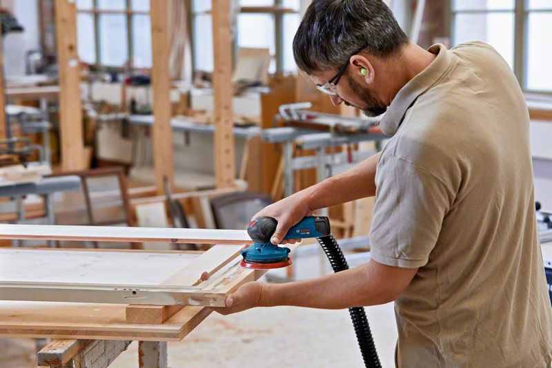 Hombre en taller lijando marco de madera con lijadora eléctrica azul, usa protección auditiva.
