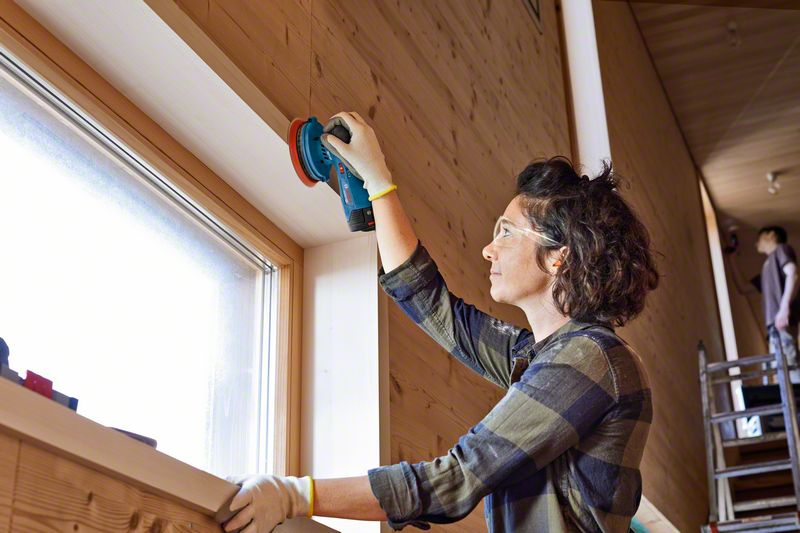 Una mujer lija una pared de madera junto a una ventana con una lijadora azul.