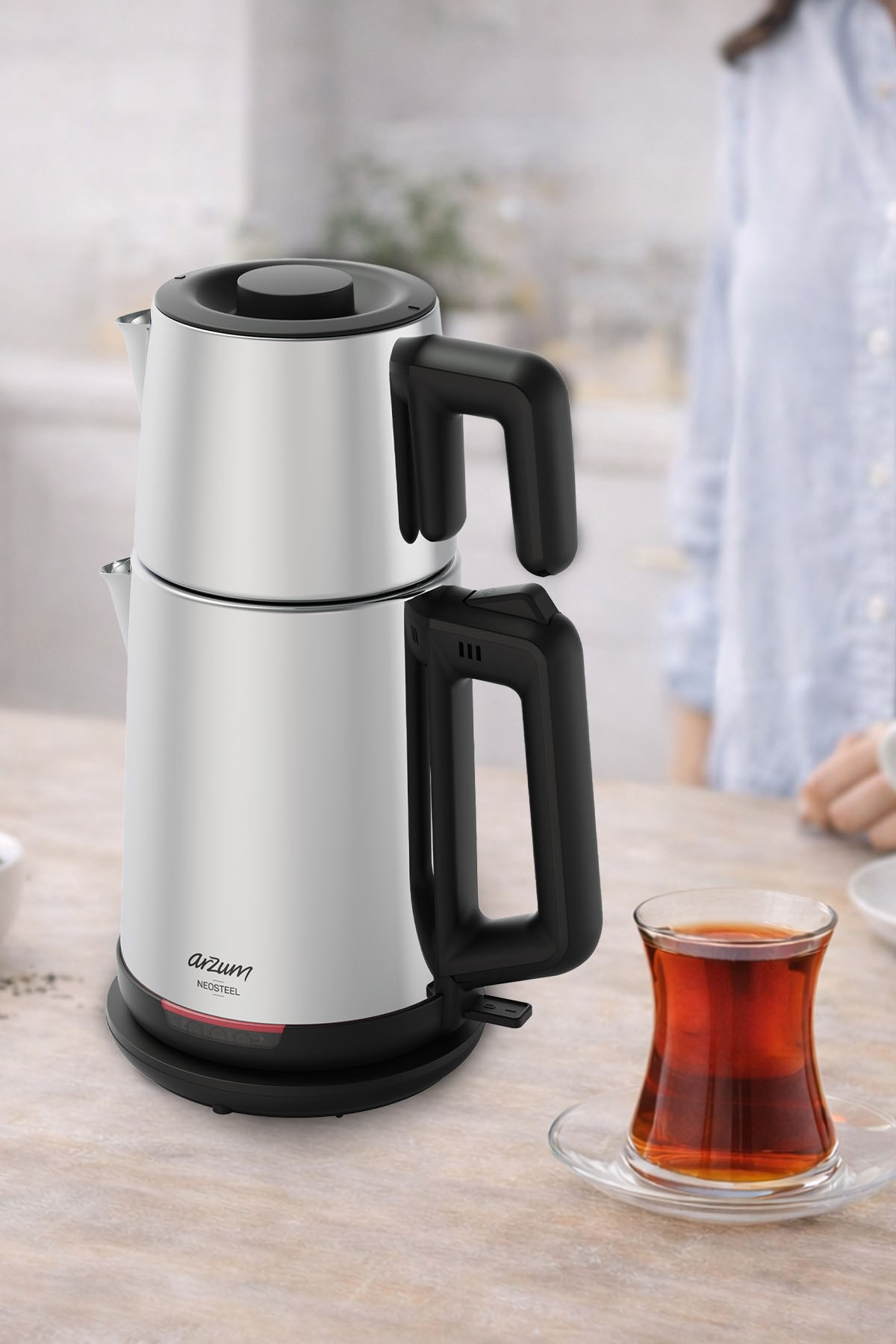 Gümüş çay makinesi, siyah kulplu, bir bardak çayın yanında.