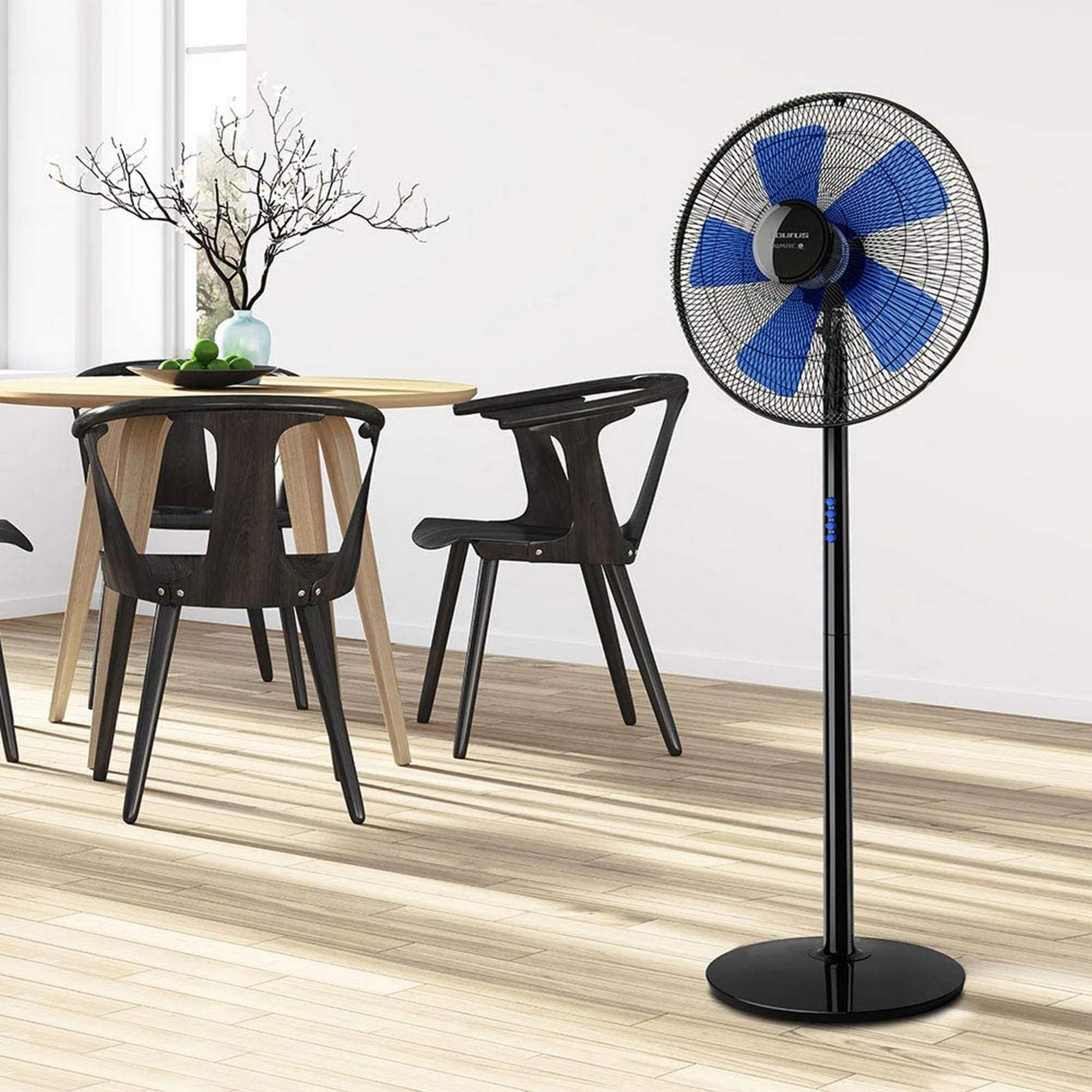 Un ventilatore a piantana nero con pale blu si trova in una stanza con tavolo e sedie.