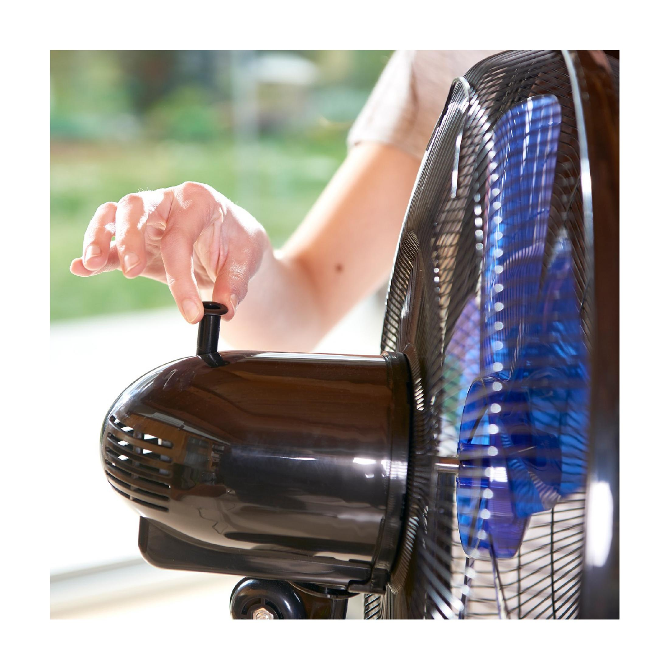 Una mano regola un ventilatore marrone, pale blu visibili.