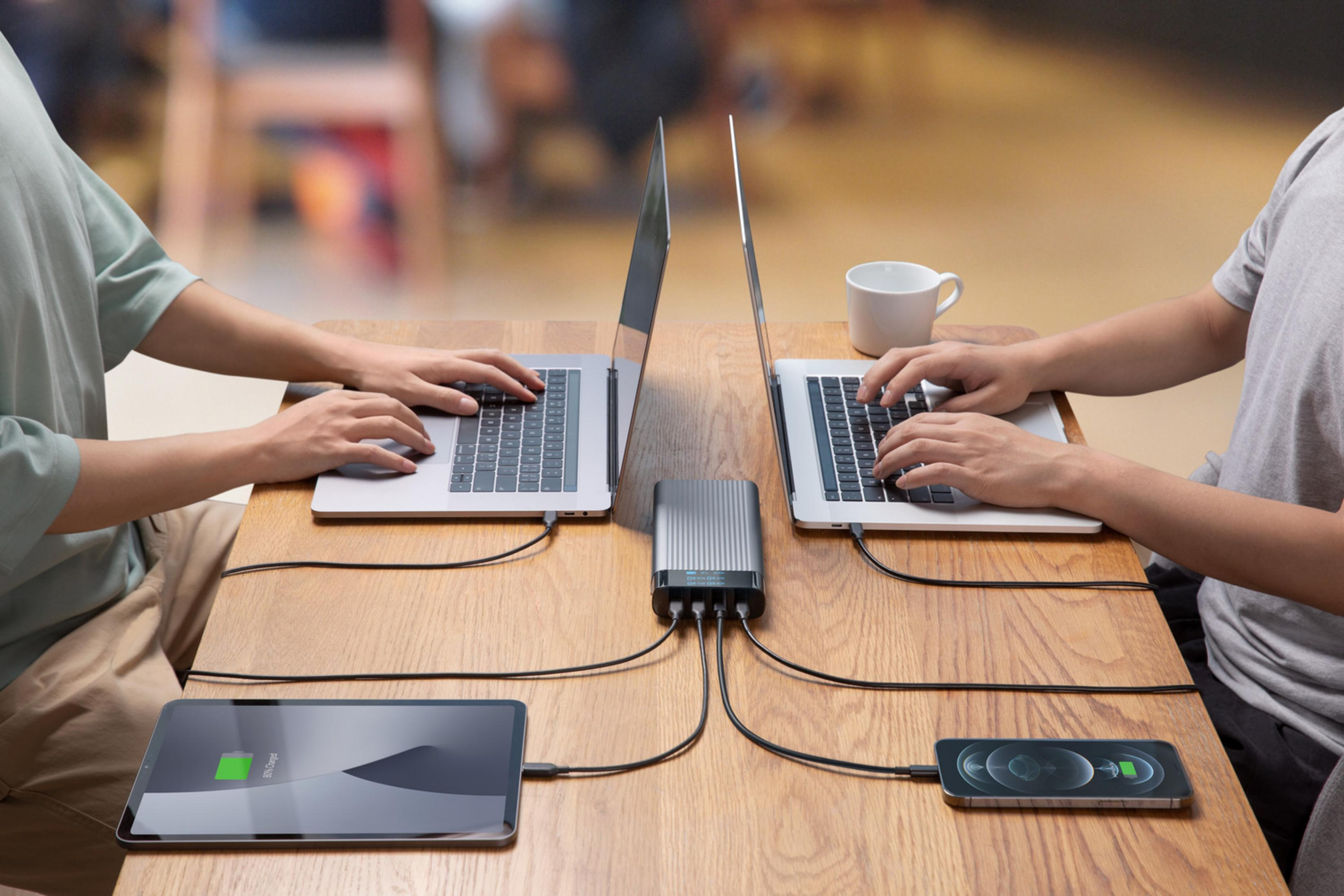 Dos personas trabajan en portátiles mientras una tablet y un móvil se cargan.