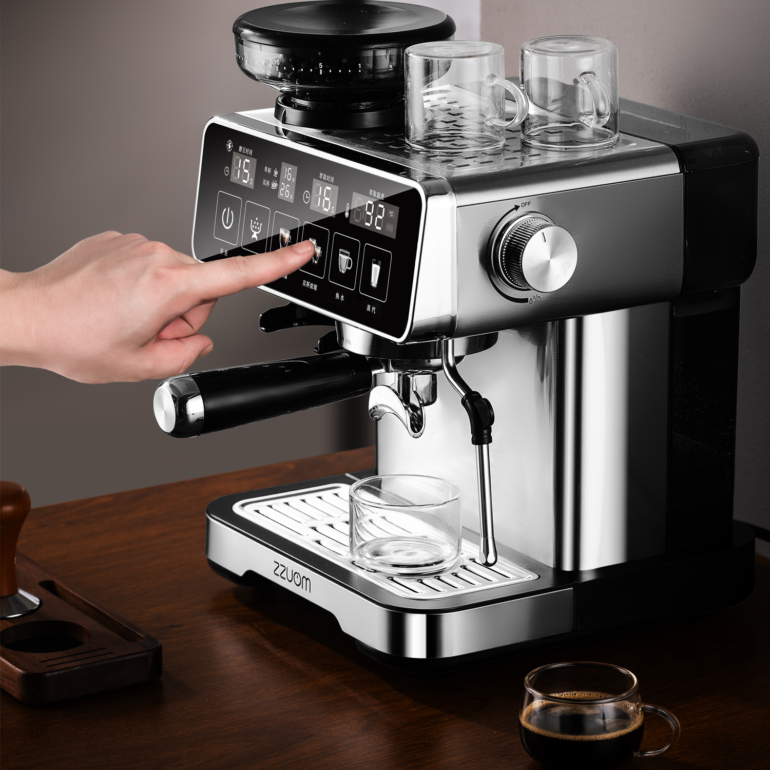Eine Hand drückt auf das Bedienfeld einer silbernen Espressomaschine. Auf der Ablage stehen Gläser.