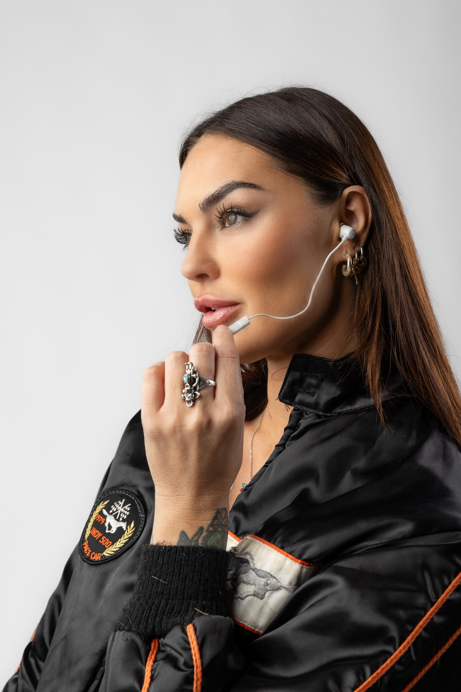 Eine Frau mit weißen Ohrhörern und auffälligem Schmuck trägt eine schwarze Jacke.
