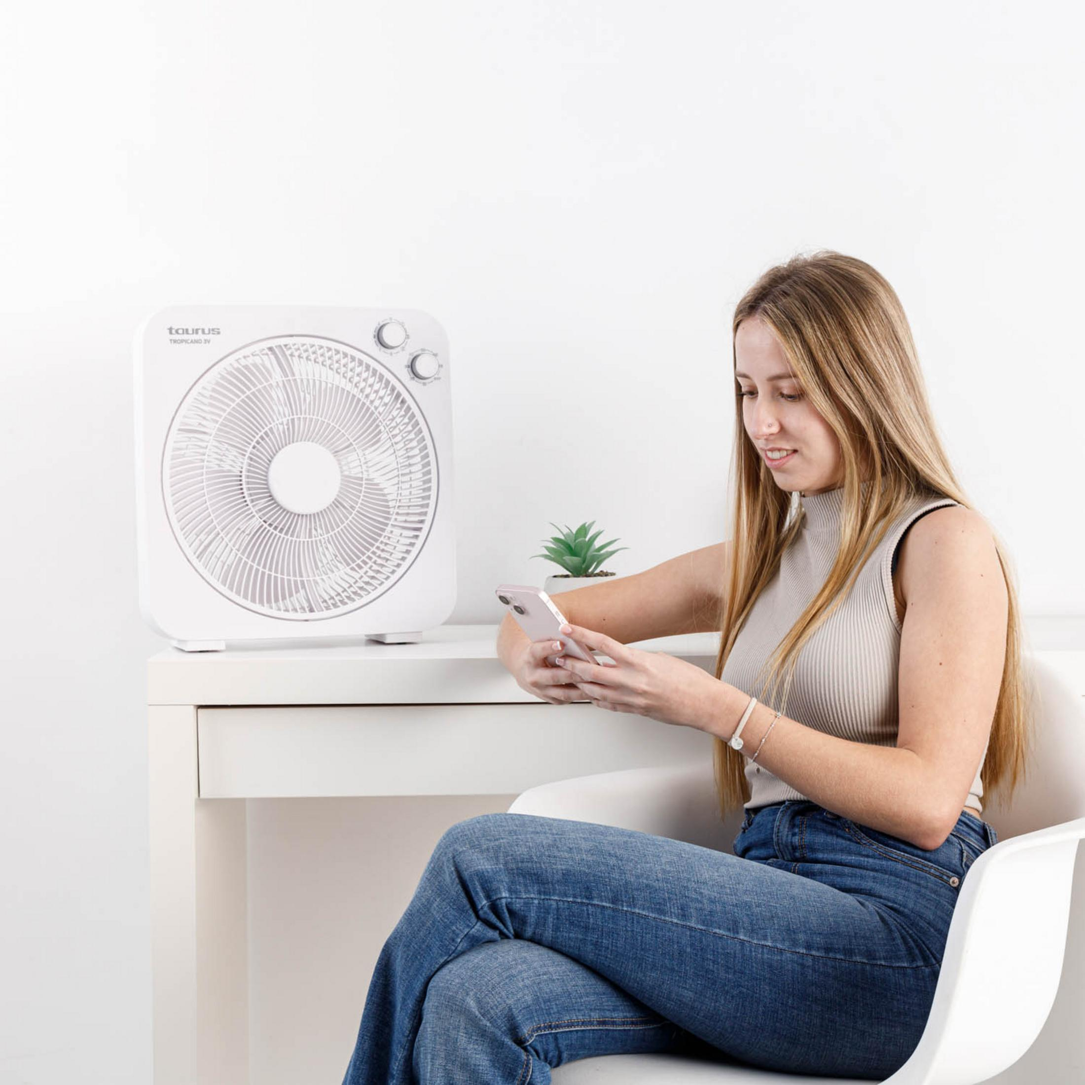 Una joven se sienta en una silla mirando su teléfono. Un ventilador está sobre un escritorio.