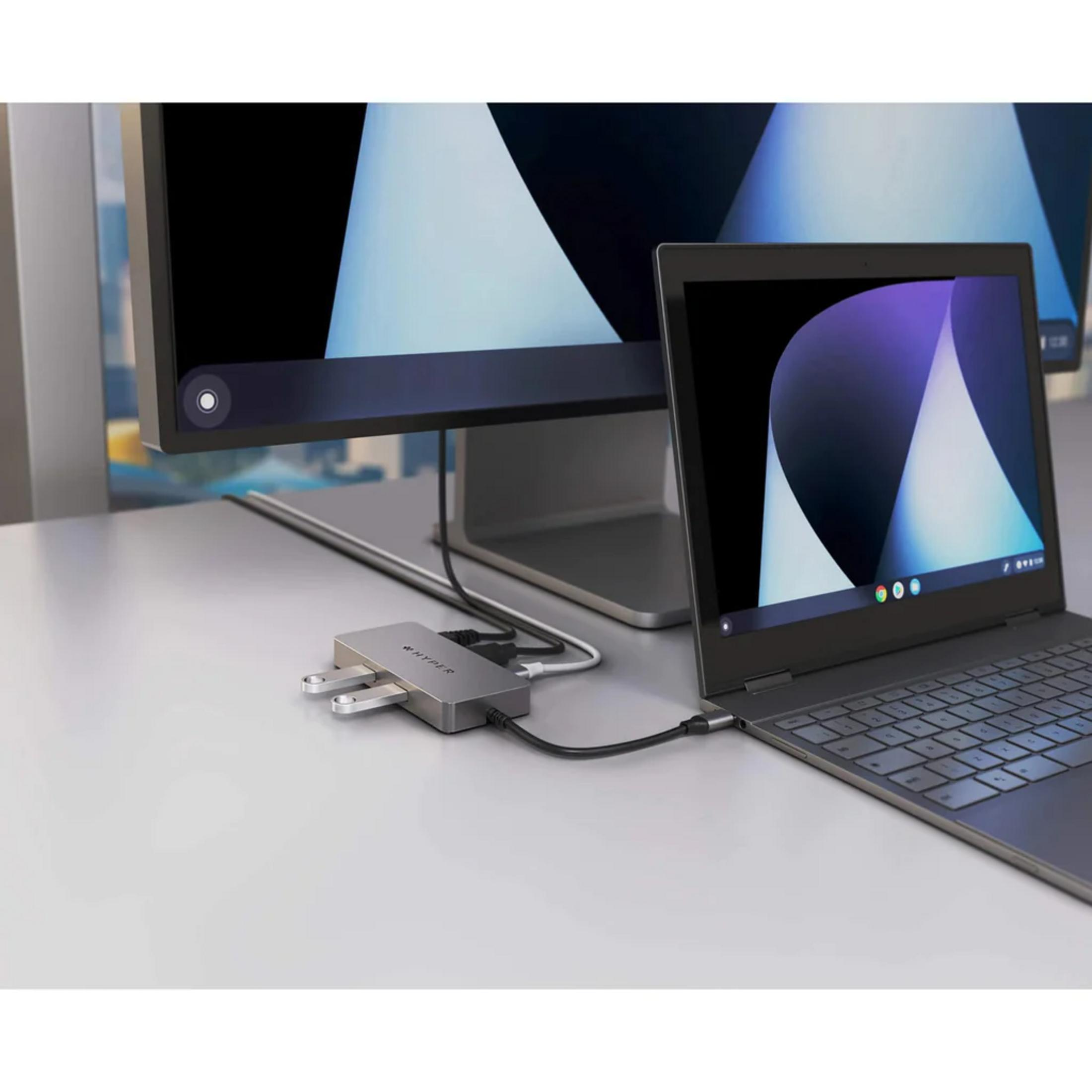Un concentrador USB-C plateado conecta un portátil y un monitor. Hay cables conectados.