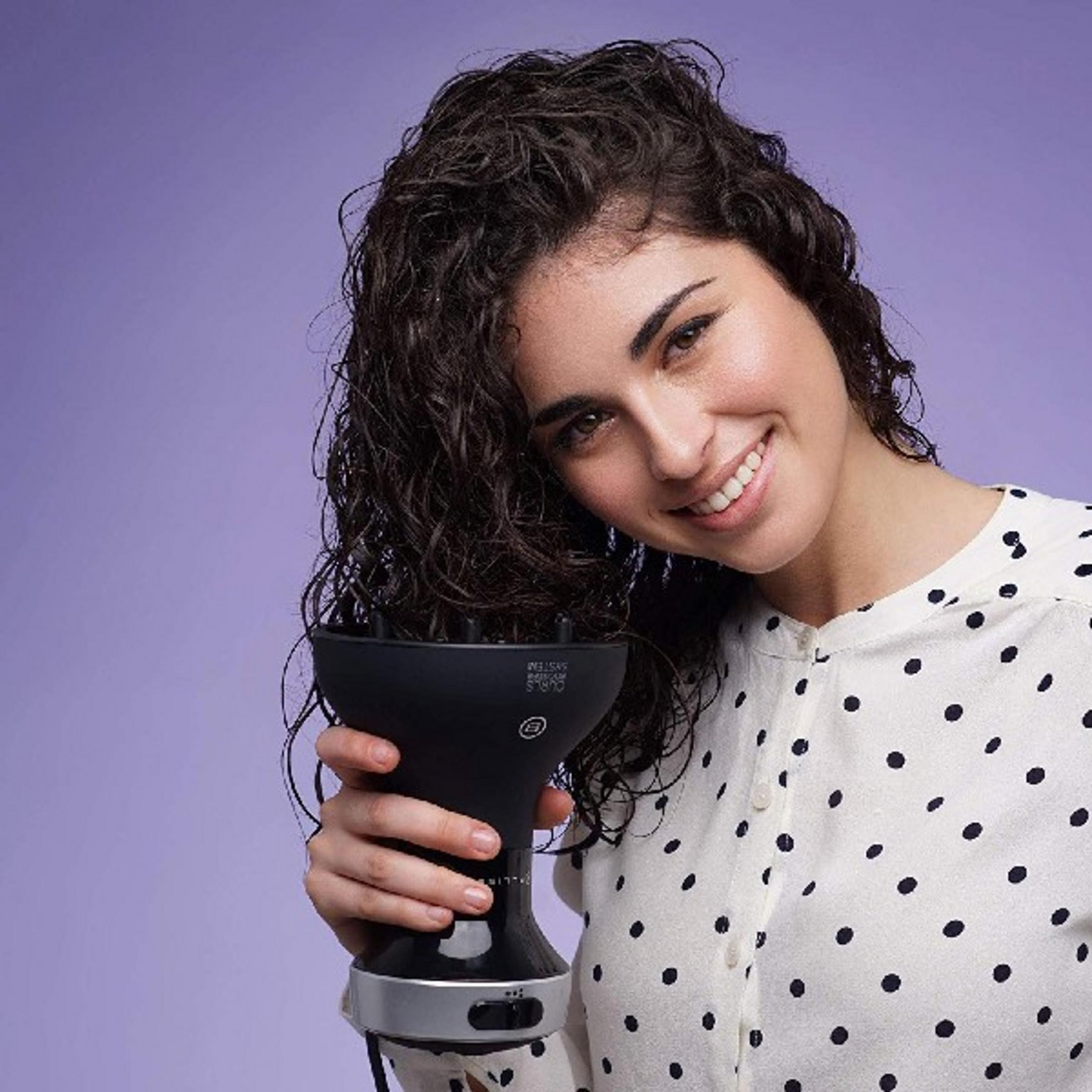 Donna con capelli ricci tiene diffusore nero su sfondo viola.