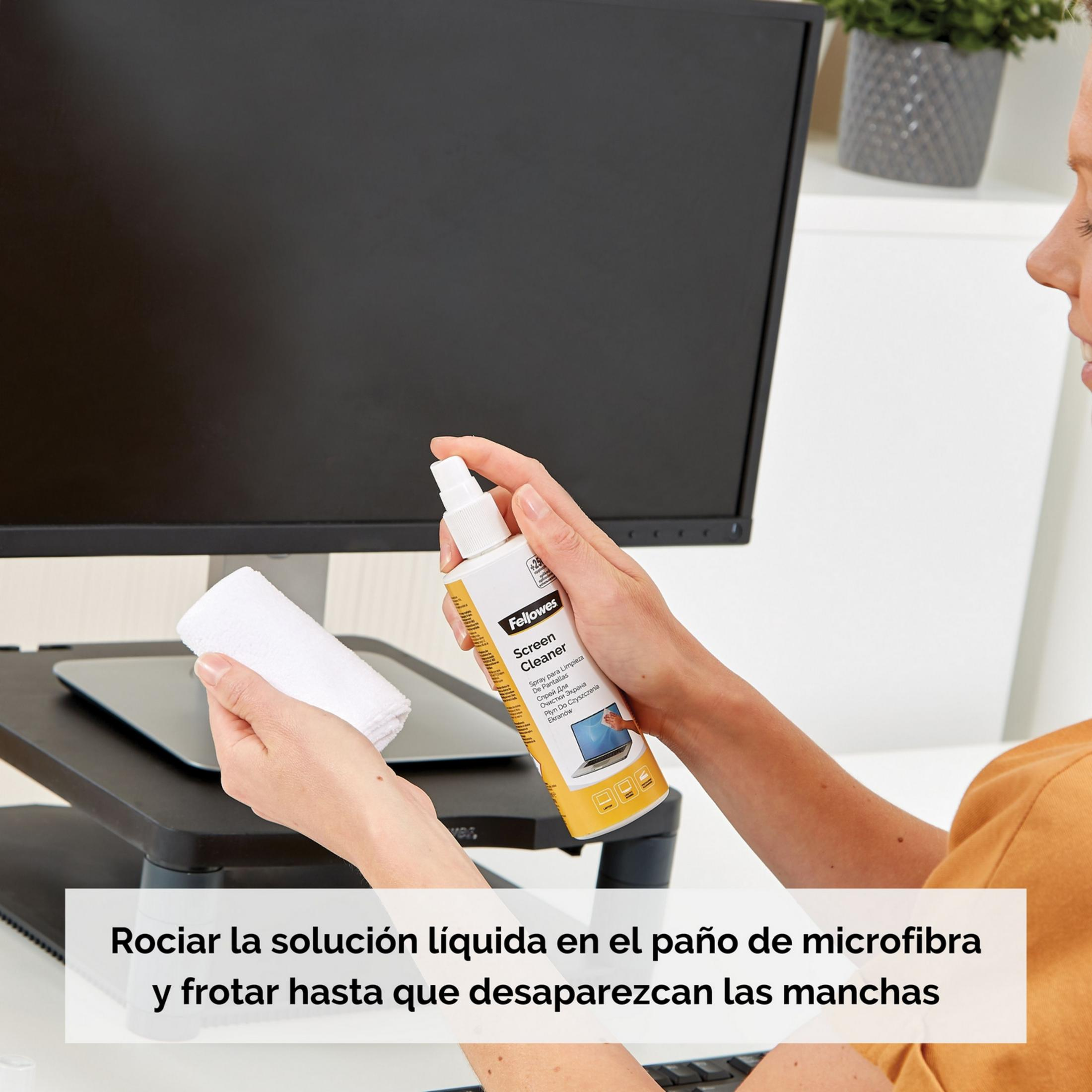 Una mano spruzza detergente su un panno in microfibra davanti a un monitor.