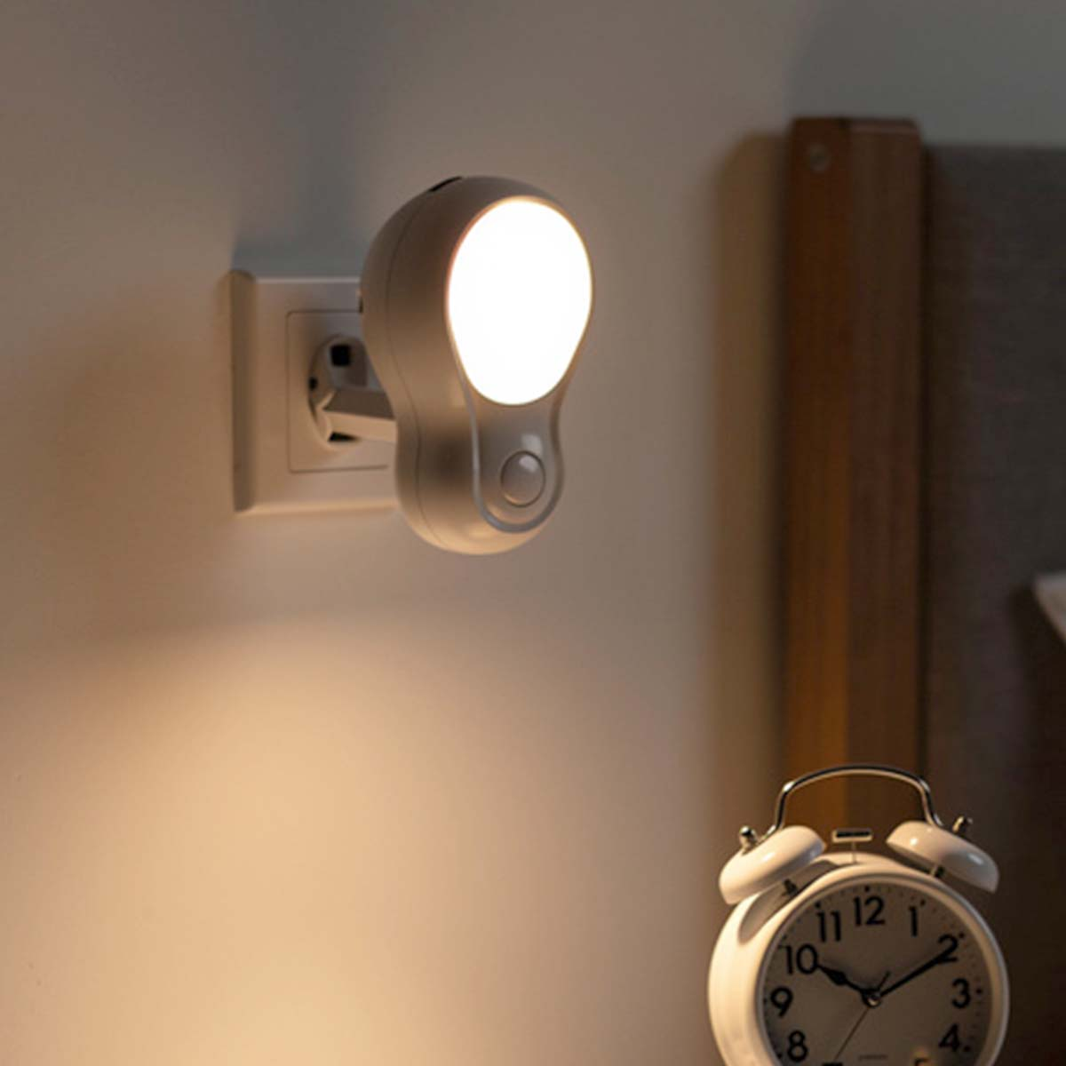 Una luz nocturna blanca con forma de bombilla se enchufa a la pared cerca de un despertador.