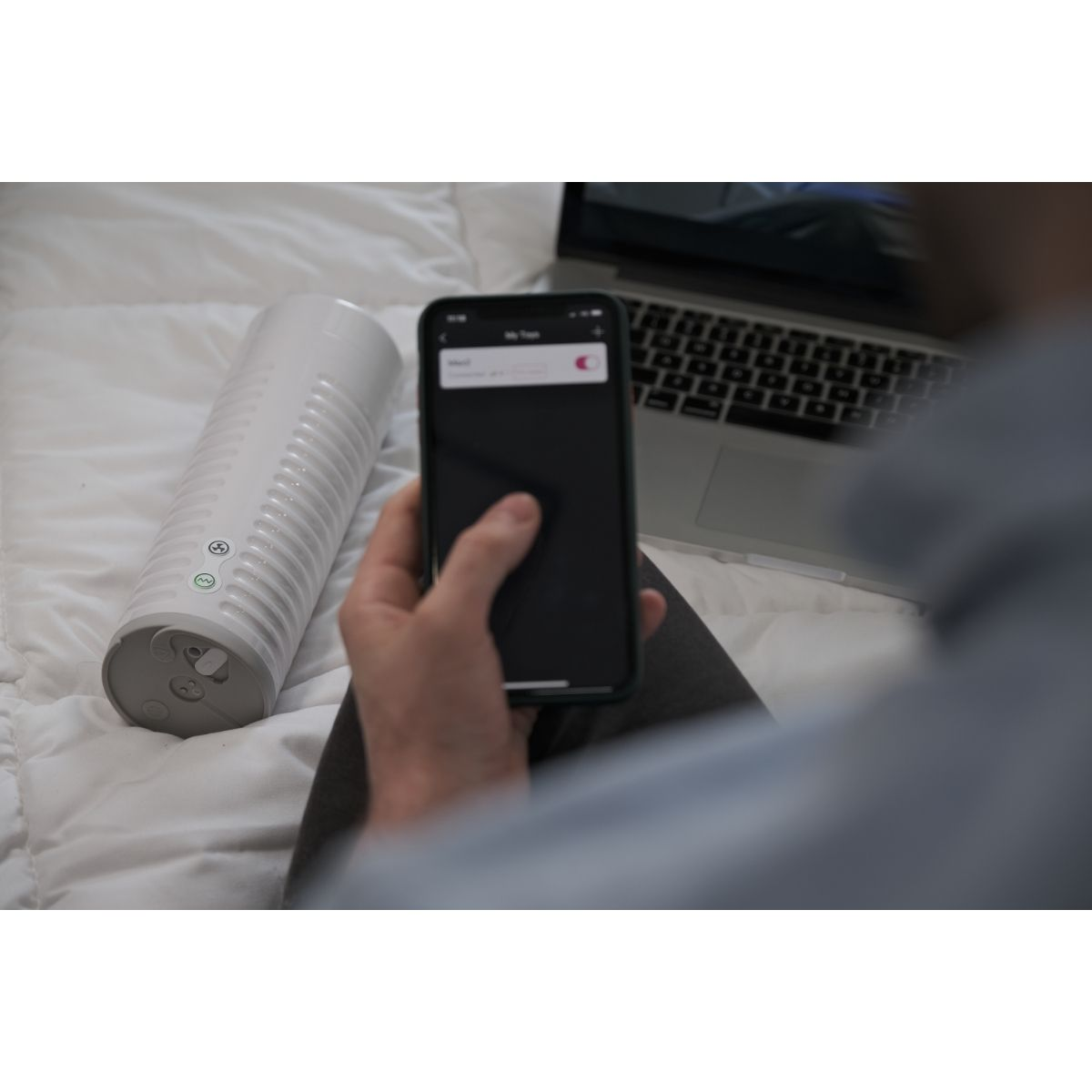 Osoba trzyma telefon, z białym cylindrycznym urządzeniem i laptopem na łóżku.