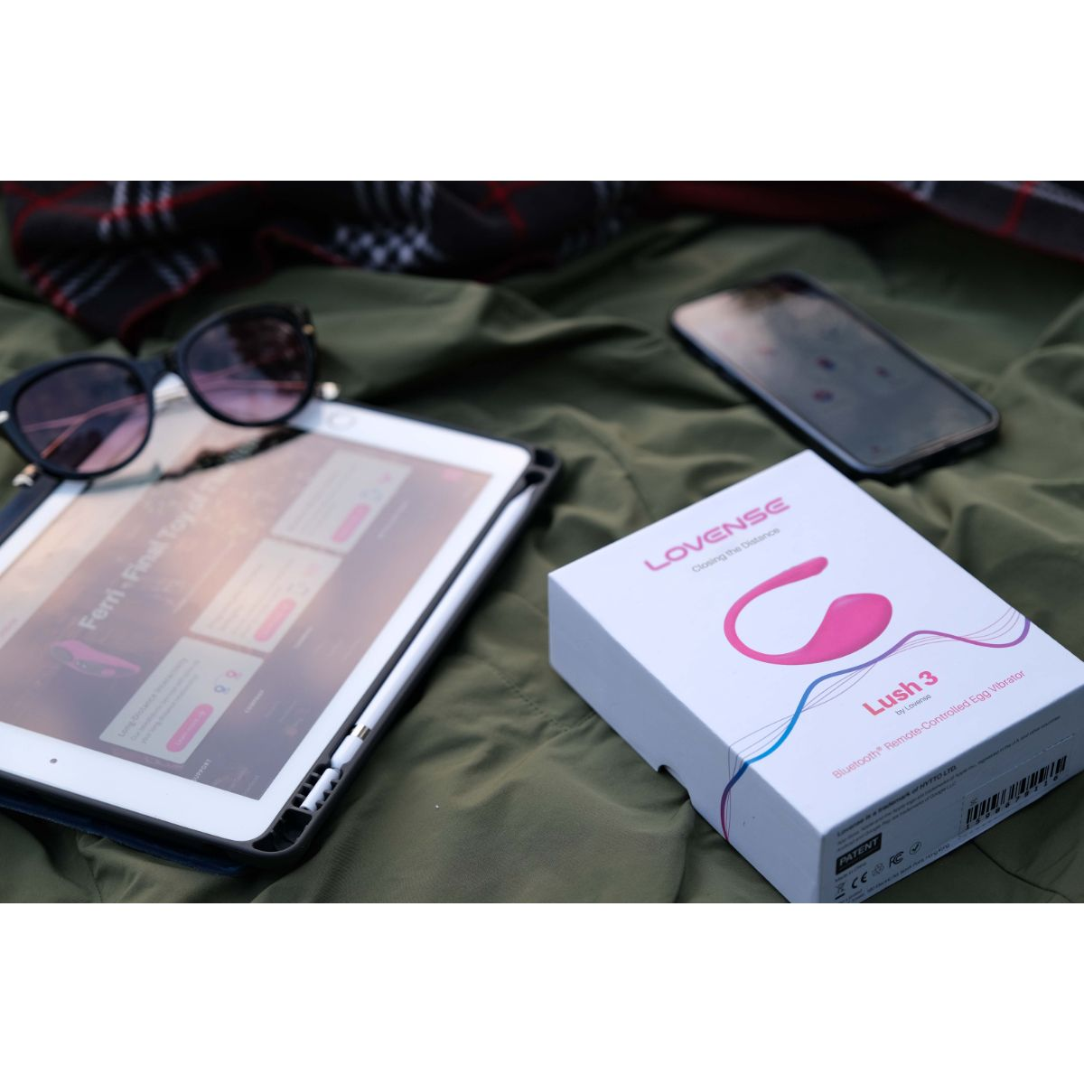 Tablet, okulary przeciwsłoneczne, smartfon i pudełko wibratora Lovense Lush 3 na tkaninie.