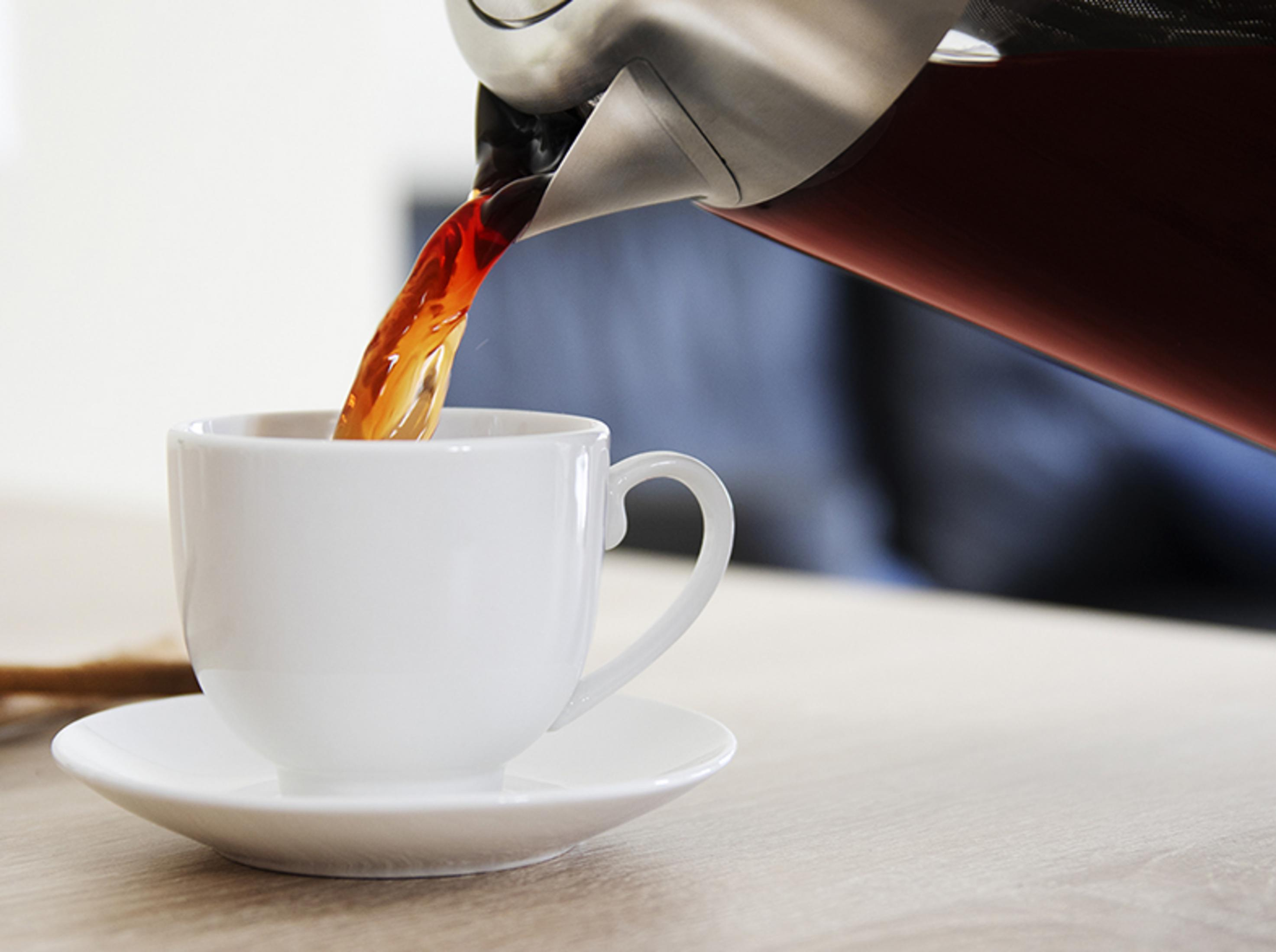 Caffè scuro versato da una caffettiera in una tazza bianca su un tavolo di legno.