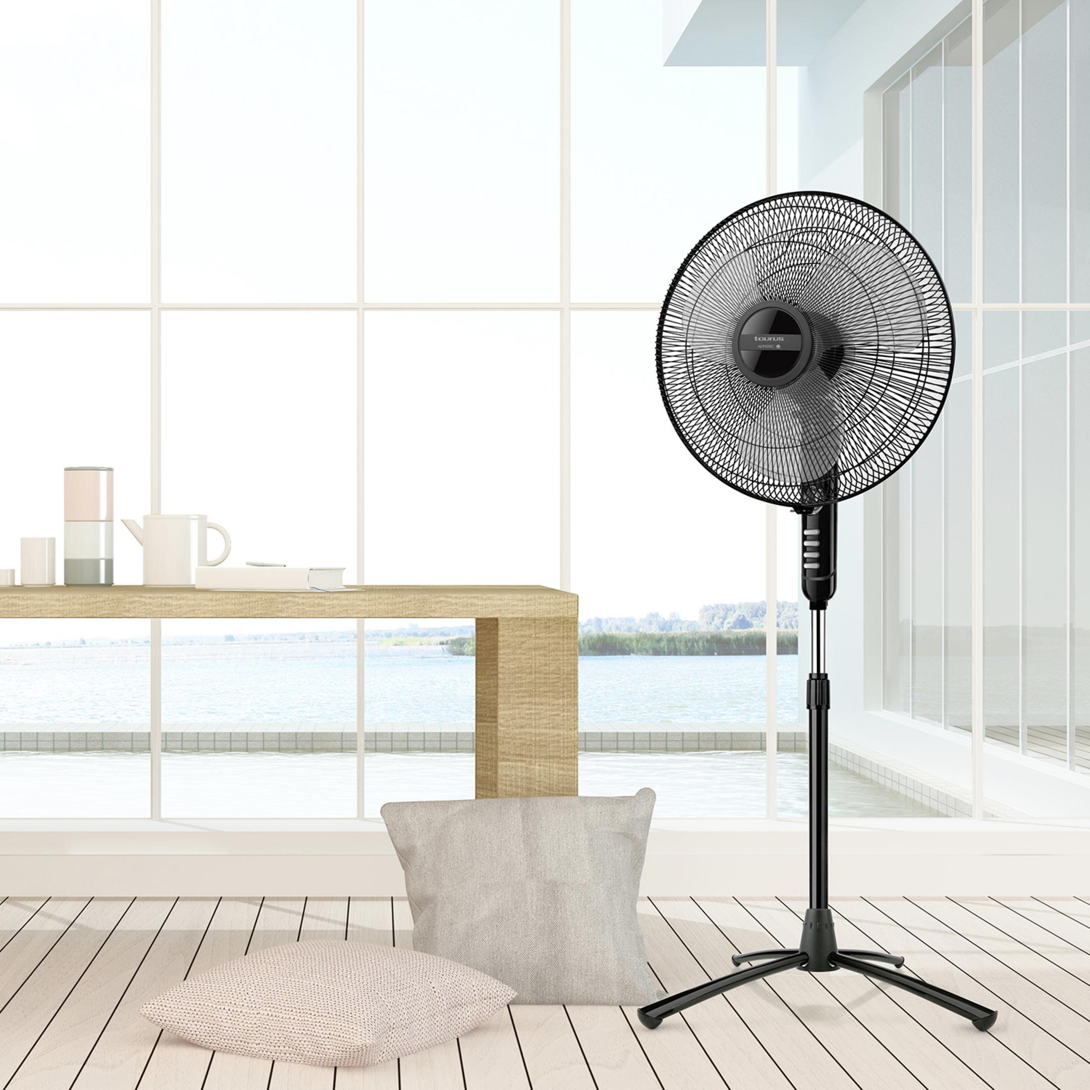Un ventilatore da terra nero si trova su un pavimento in legno di fronte a una grande finestra con vista sull'acqua.