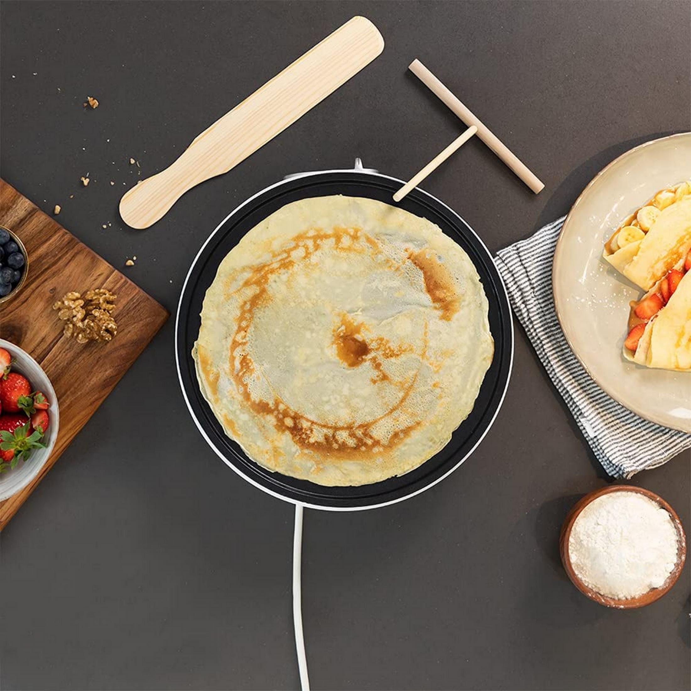 Una grande crêpe cuoce su una piastra elettrica con ingredienti nelle vicinanze.