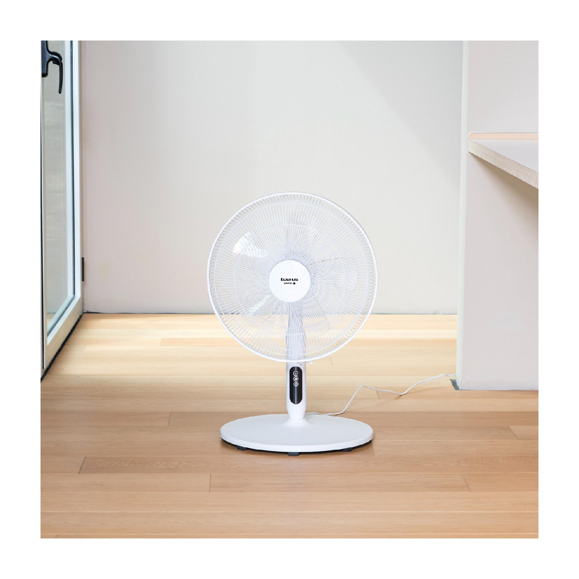 Un ventilatore da terra bianco si trova su un pavimento in legno di fronte a un muro chiaro.