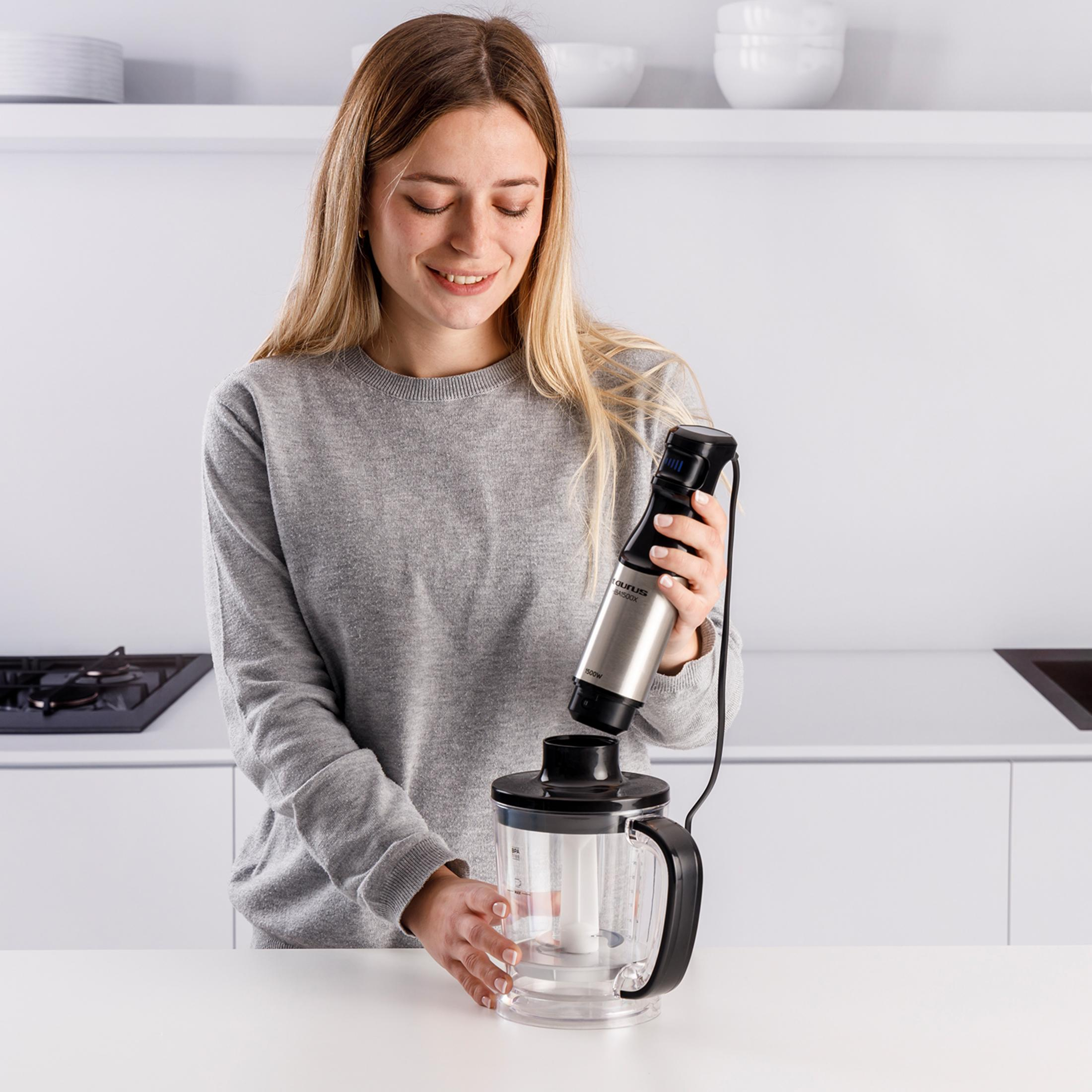 Una donna con i capelli biondi tiene un frullatore a immersione sopra un misurino.