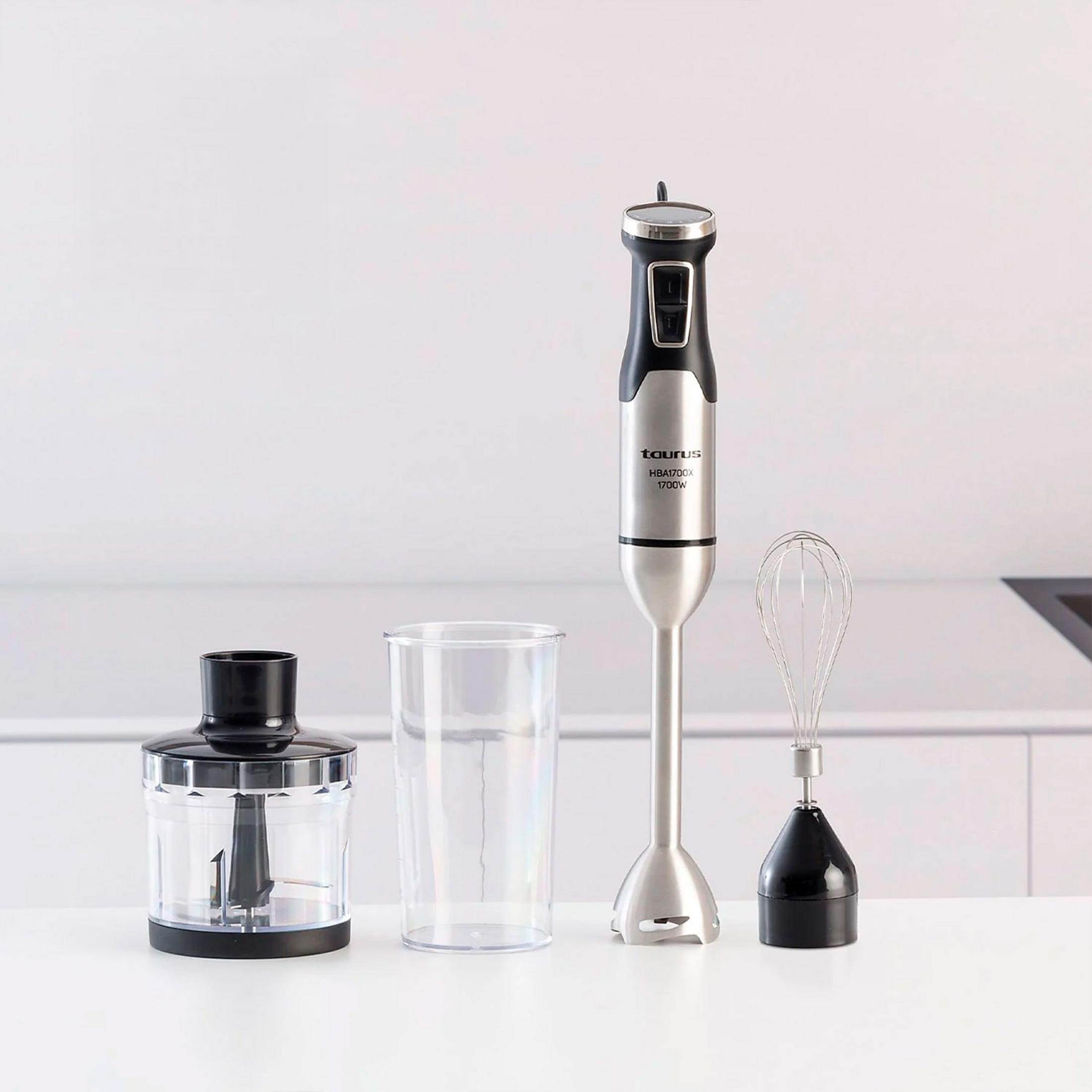 Un frullatore a immersione con accessori: bicchiere, tritatutto e frusta su tavolo bianco.