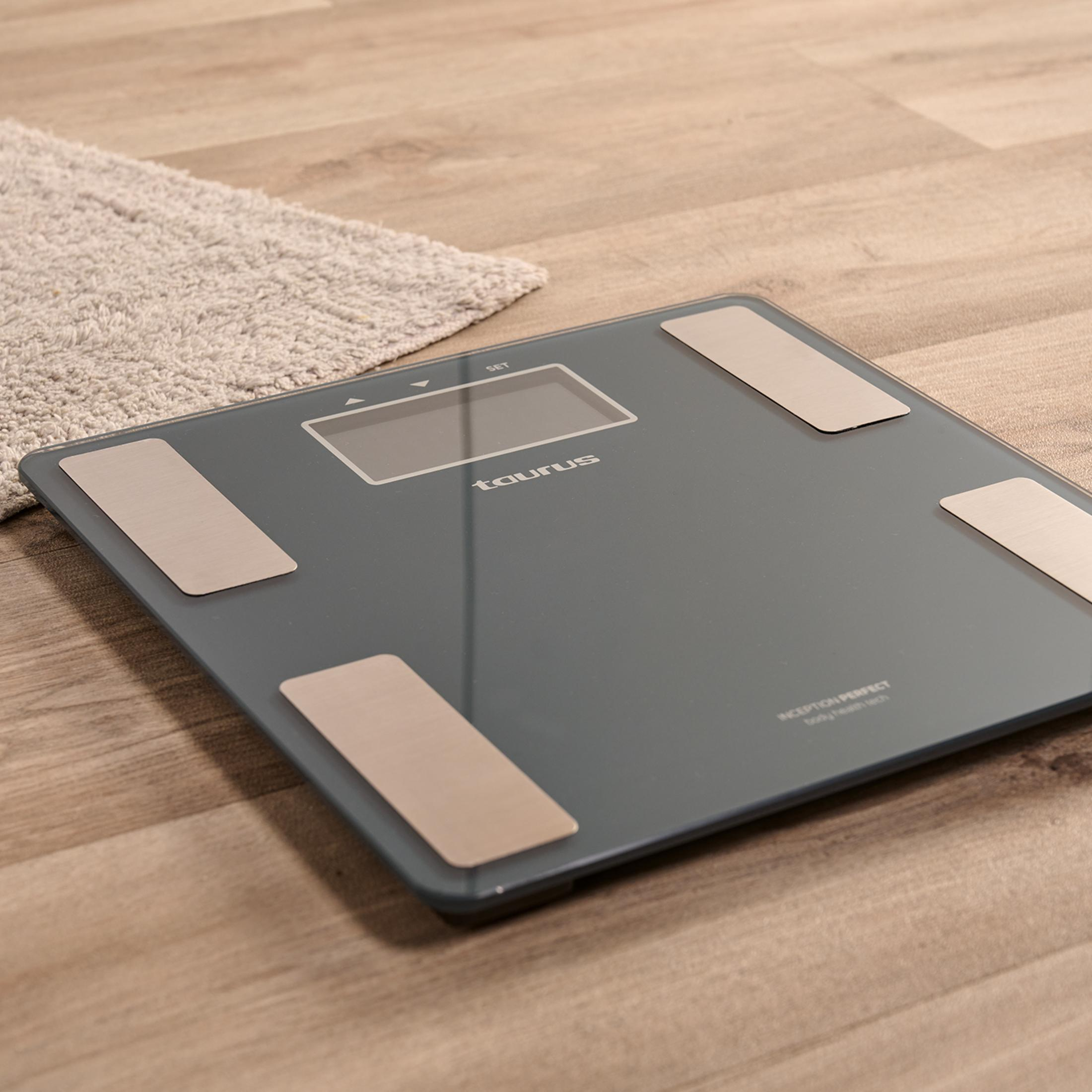Een grijze digitale weegschaal op een houten vloer met een klein vloerkleed ernaast.