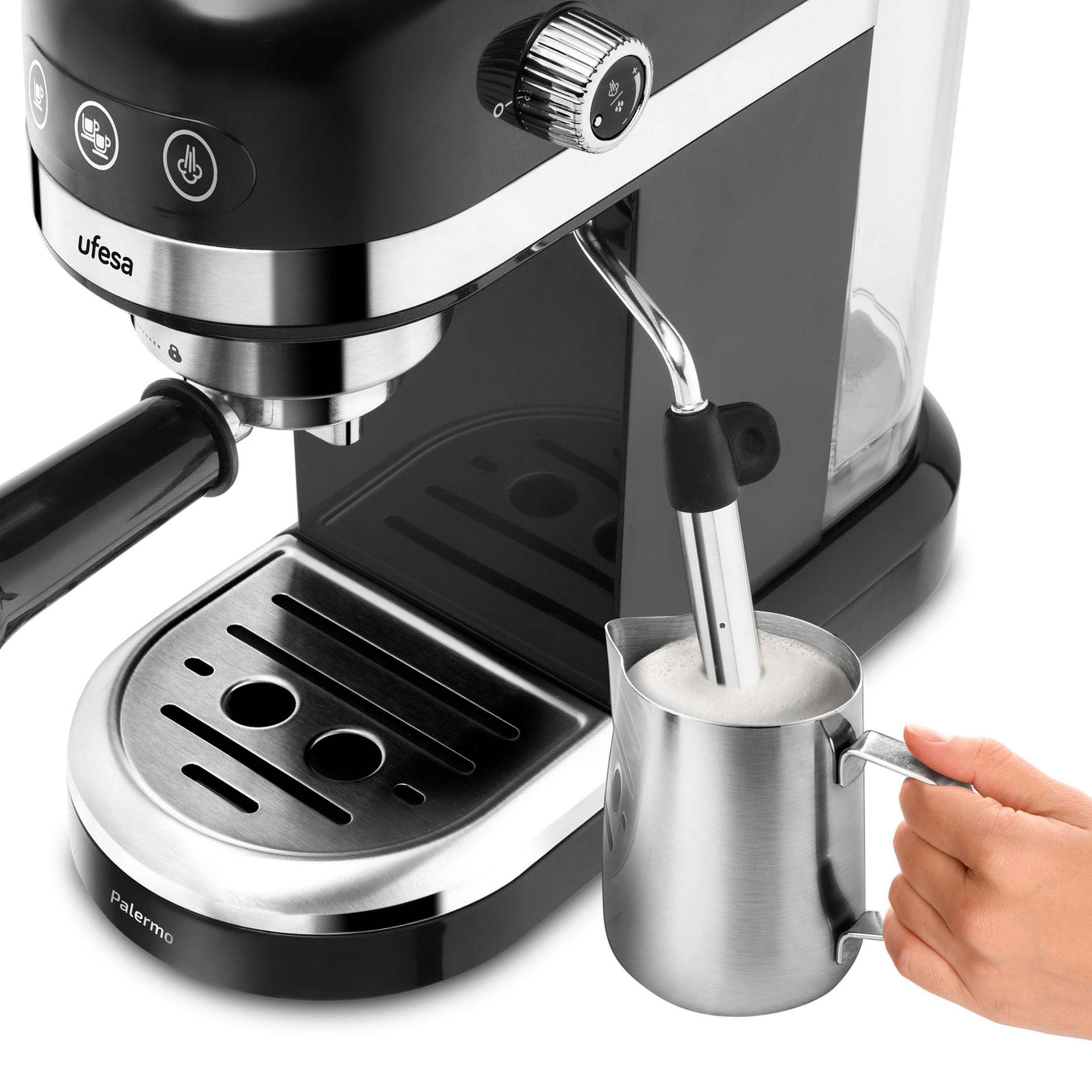Una mano tiene una lattiera per montare il latte accanto a una macchina per espresso nera.