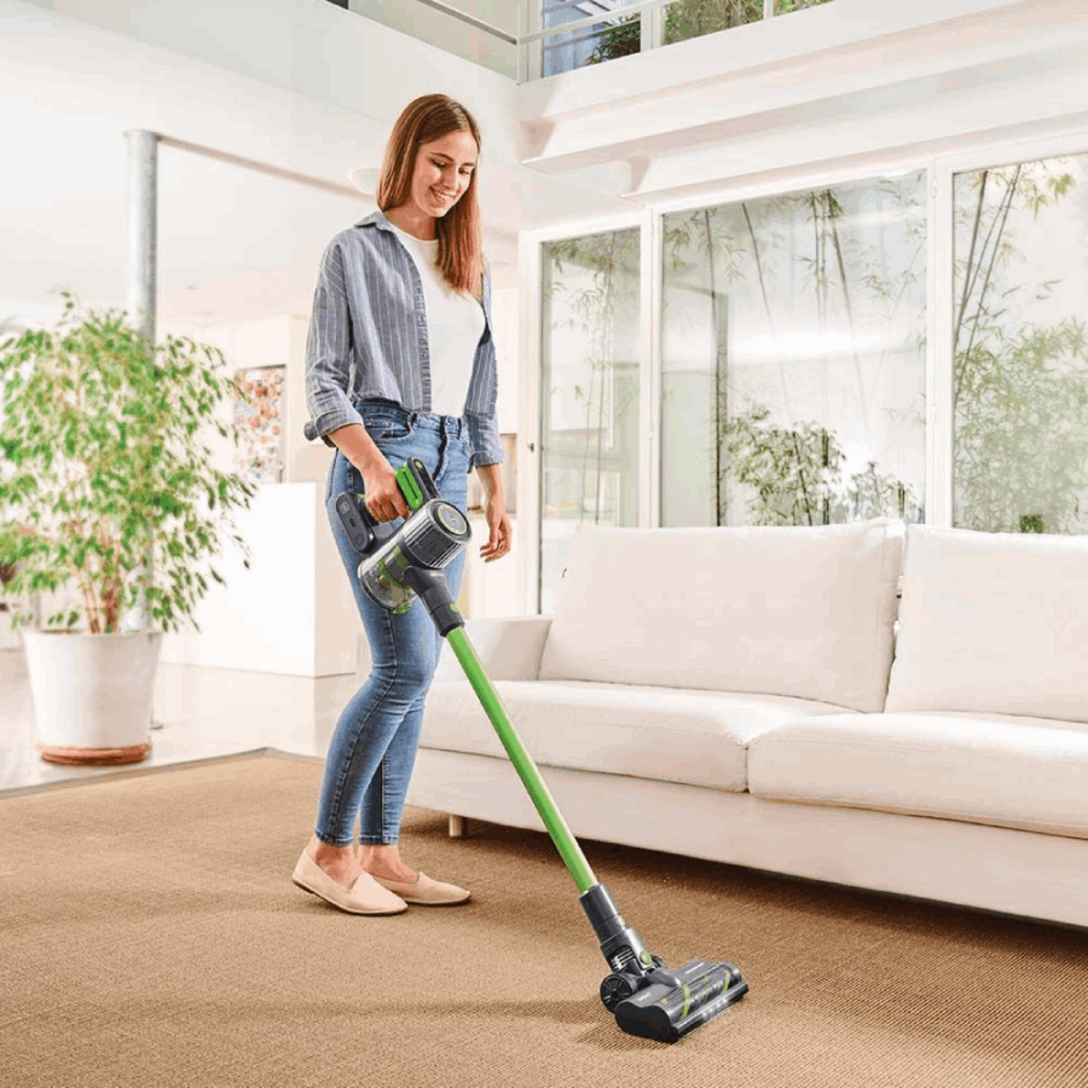 Una mujer aspira una alfombra con una aspiradora inalámbrica verde en una sala de estar.