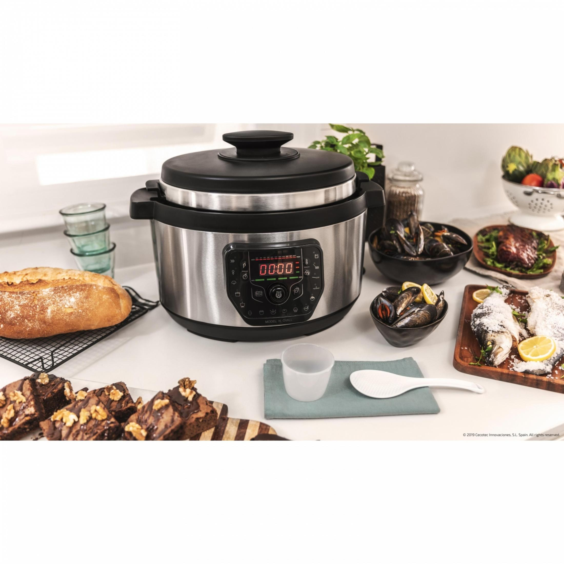 Una olla a presión eléctrica plateada en un mostrador con pan, pastel y platos de mariscos.
