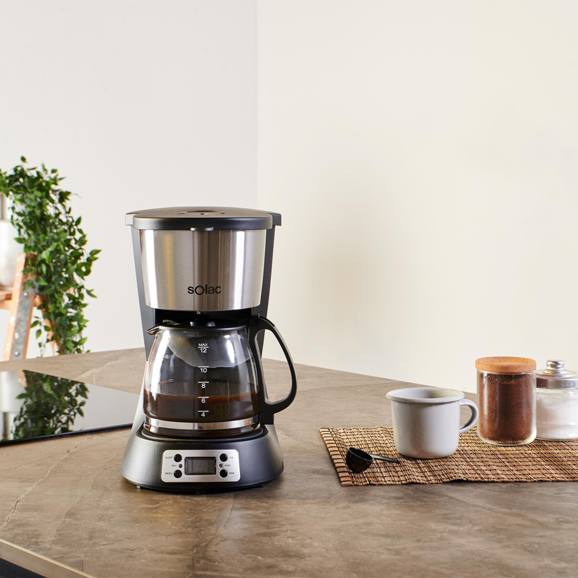 Cafetera con jarra de cristal, taza, café molido y azúcar sobre una encimera.