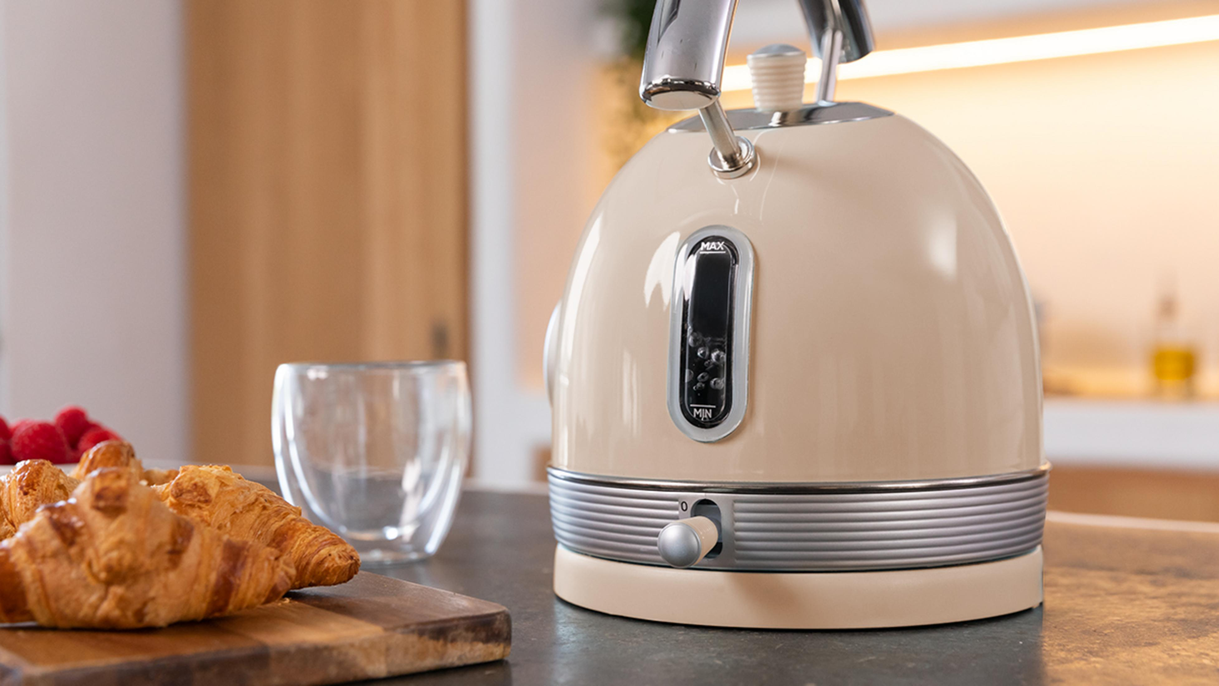 Ein beiger Wasserkocher mit Crombesatz steht auf einer Arbeitsplatte neben Croissants und einem Glas.