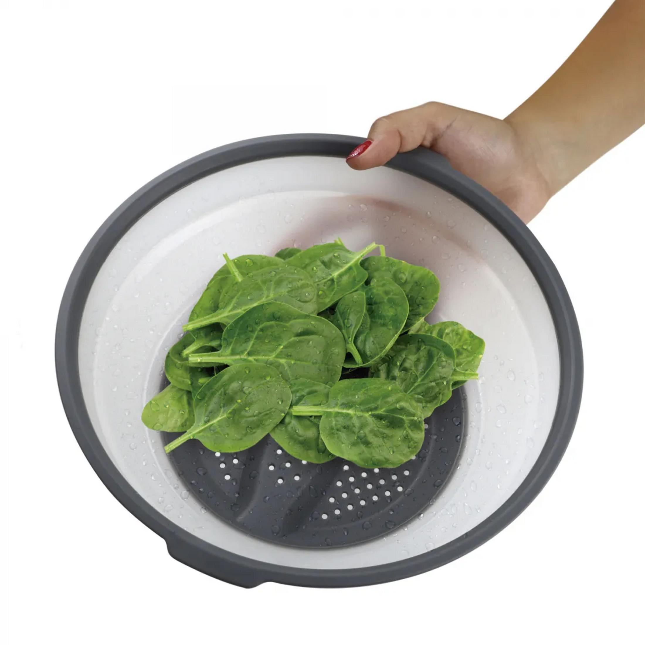 Una mano sostiene un colador gris y blanco con espinacas frescas y gotas de agua.