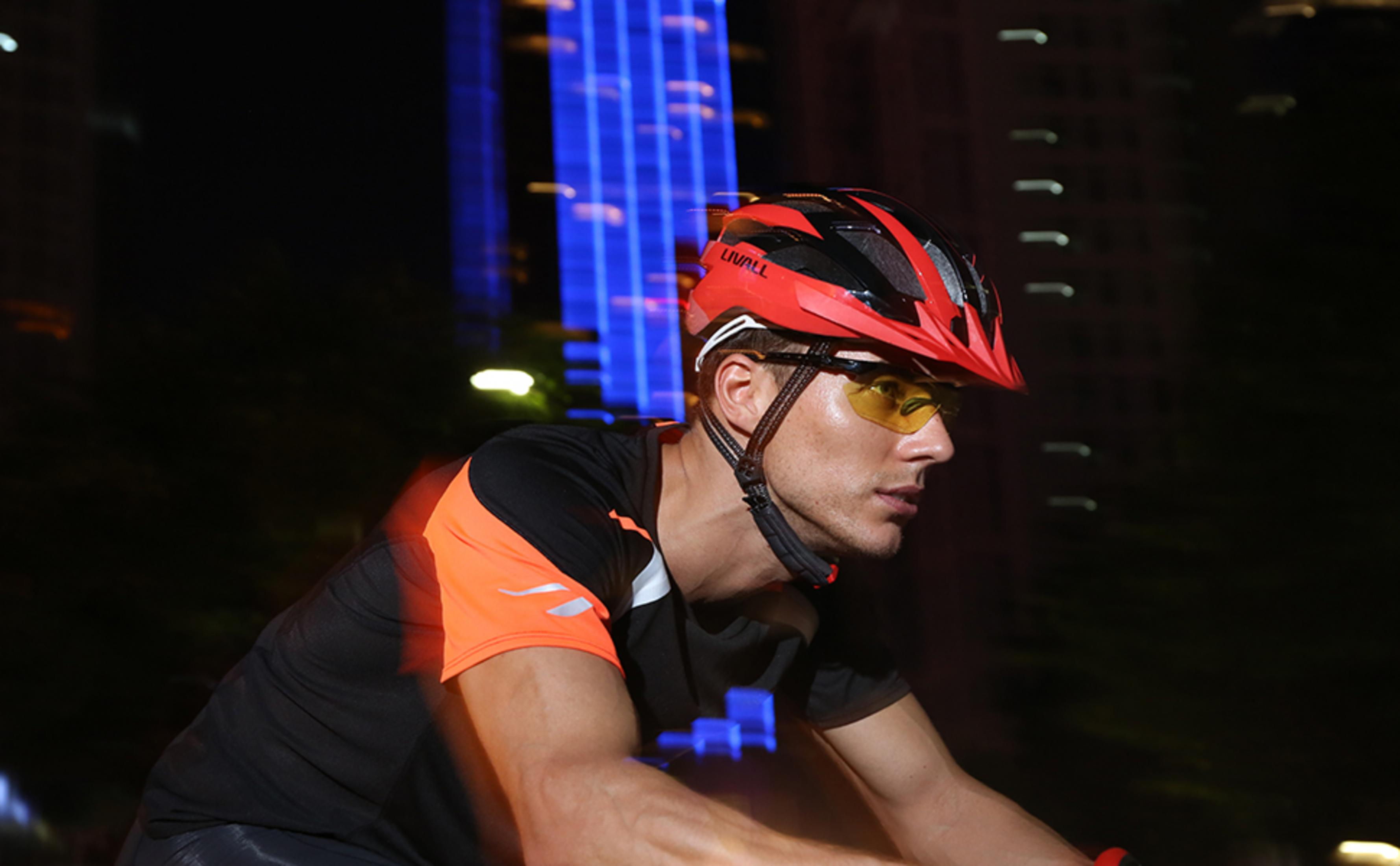 Un uomo con casco rosso e occhiali gialli va in bicicletta di notte.